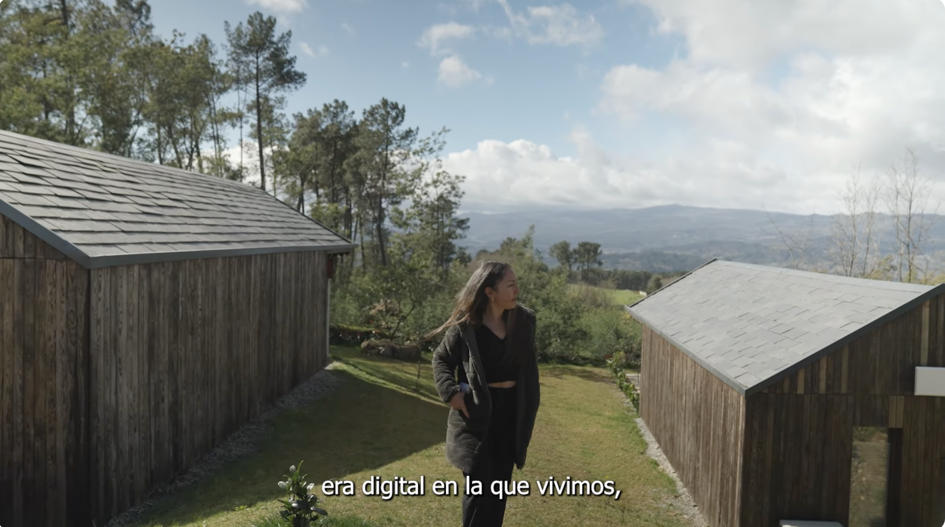 Persona caminando entre dos construcciones de madera en un entorno rural con césped, árboles y montañas al fondo, bajo un cielo parcialmente nublado. En la parte inferior aparece el texto ‘era digital en la que vivimos’.