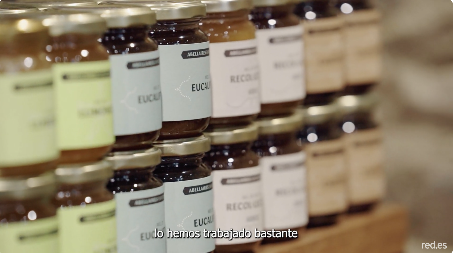 Fila de tarros de miel artesanal con etiquetas que indican variedades como eucalipto y otras, colocados en una estantería de madera. En la esquina inferior aparece el texto ‘red.es’ y subtítulos parciales en español.