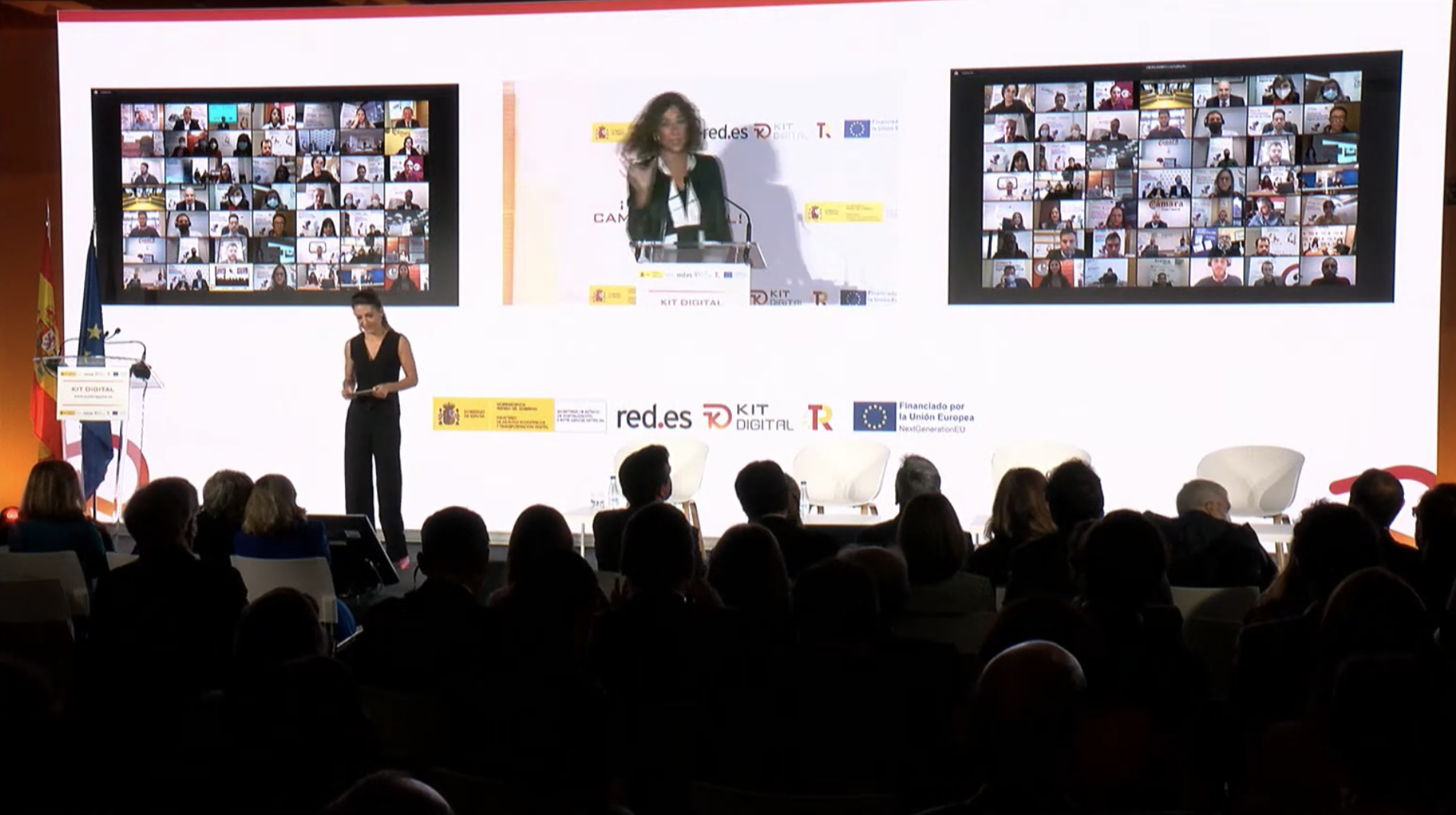 Evento presencial sobre el programa Kit Digital con público sentado frente a un escenario, donde una persona habla en un atril y otra está de pie. En la pantalla se muestran dos mosaicos con participantes conectados por videoconferencia y logotipos institucionales como red.es, Kit Digital y la Unión Europea.